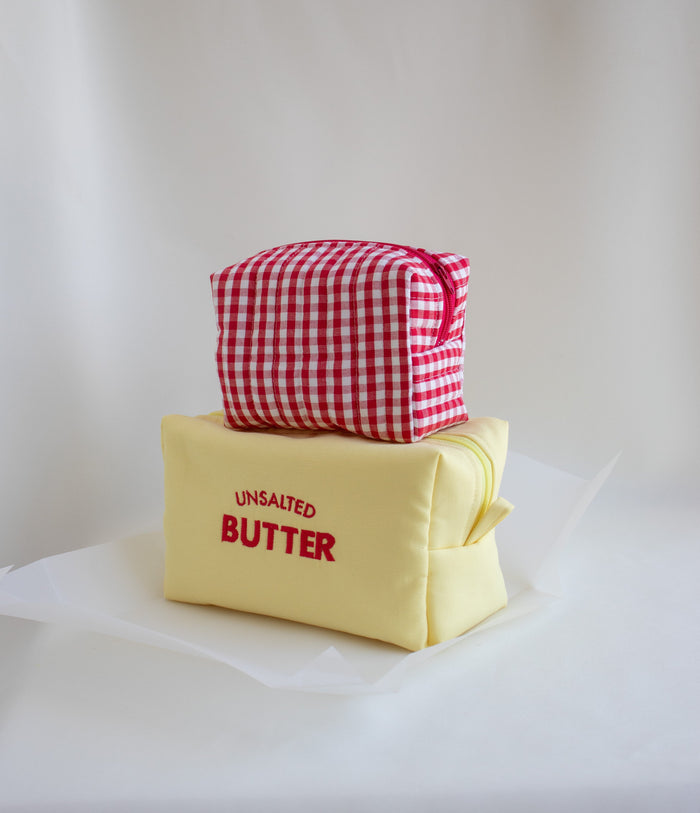 Two fabric cosmetic pouches stacked on a white surface against a white draped backdrop. The bottom pouch is pale yellow with red text reading 'UNSALTED BUTTER,' and the top pouch is red-and-white gingham with a red zipper pull.