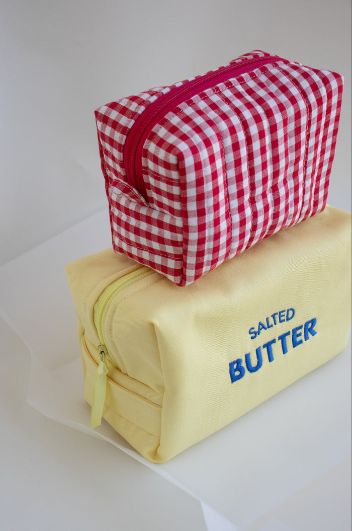 Two zippered cosmetic pouches stacked on a white surface. The top pouch is red and white gingham with a pink zipper, and the bottom pouch is pale yellow with blue text reading 'SALED BUTTER'.