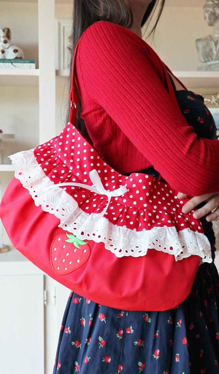 A red shoulder bag with white polka dot fabric, white lace ruffles, and a strawberry applique, carried by a person wearing a red ribbed sweater and a navy blue strawberry-print skirt.
