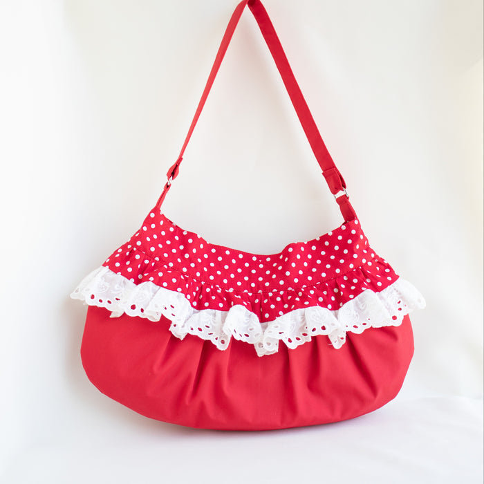 A red shoulder bag with a red and white polka dot fabric top, white eyelet lace ruffles, and a red shoulder strap, shown against a white background.